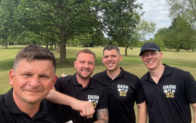 4 men in black polos smiling with trees in golf course in background