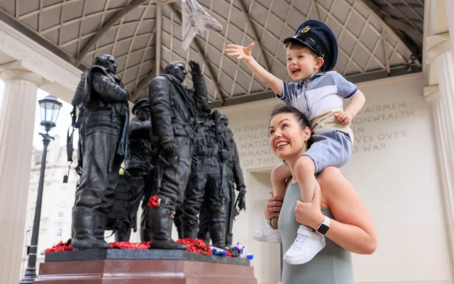 A child flying a tin foil aeroplane on his mum's shoulders outside the Bomber Command Memorial