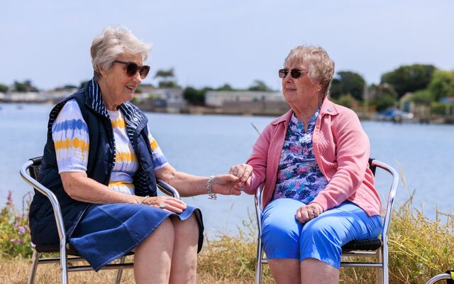 Maureen and Brenda holding hands sitting down in front of lake