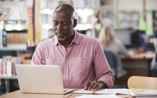 A stock image of a man sat in a library at a table, using a laptop with pens and papers on the table.