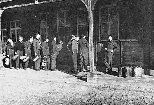 A group of PoWs hold various jugs and pots as they queue for their water ration outside a large hut at Stalag Luft III.