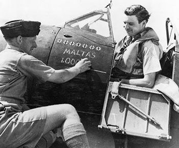 Sqn Ldr JJ Lynch, Officer Commanding 249 Squadron, sits in the cockpit of his Supermarine Spitfire Vc at Krendi, Malta, as an airman chalks "Malta's 1,000th" below his persoanl victory tally. (UK Crown Copyright / MOD. Courtesy of Air Historical Branch)