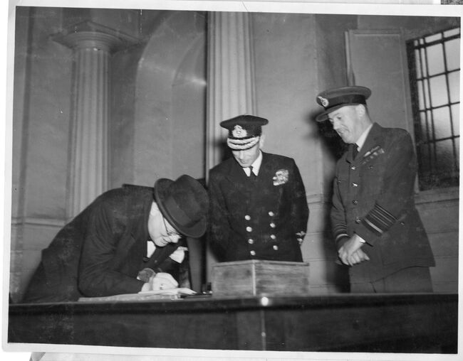 The Prime Minister, The Rt Hon Winston Churchill, signs the 'visitors' book at AEAF Headquarters during his visit there with the King. Air Chief Marshal Sir Trafford Leigh-Mallory is on the right.