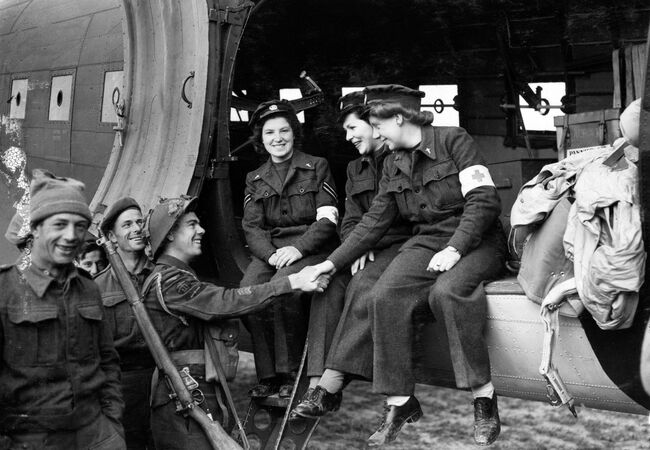 The first three nursing orderies of the Women's Auxiliary Air Force to fly to France are greeted on their arrival in Normandy by Leading Aircraftman P McGreech, a servicing commando on, 13 June 1944. From left to right, the WAAFs are: Corporal Lydia Alford and Leading Aircraftwomen Myra Roberts and Edna Birbeck.