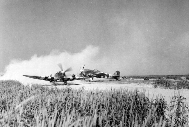 A Supermarine Spitfire IX - fitted with a 45-gallon slipper fuel tank - raises the dust as it taxies past a Hawker Typhoon Ib of 181 Squadron at B2/Bazenville, Normandy, on 17 June 1944. On the port wing can be seen a member of the groundcrew operating as a 'spotter' for the pilot to avoid holes and obstacles at the temporary airstrip.