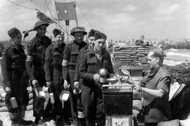Airmen of 107 Beach Flight, queue up for a meal at their cookhouse on a Normandy beach, 22 June 1944. They are (left to right): Leading Aircraftsman L Sexton of Chester, Corporal F Kirby of Edgbaston, Birmingham, Sergeant D Howells of Worthing, Corporal J Holland of St Helens, Flight-Sergeant JE Gould of Liverpool and Leading Aircraftsman G Harvey of Brighton. Serving the food is the cook, Leading Aircraftsman W Spackman of Walthamstow, London.
