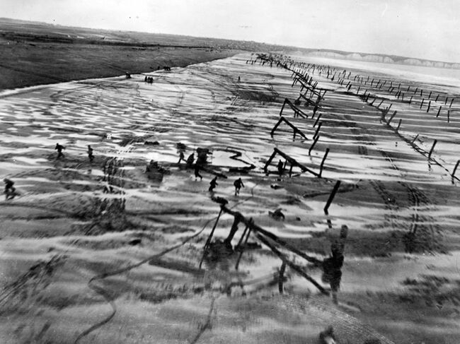 Low level photograph showing German soldiers attempting to build defences on a beach in Normandy running for cover as the reece aircraft streaks past at low level, catching them unprepared.