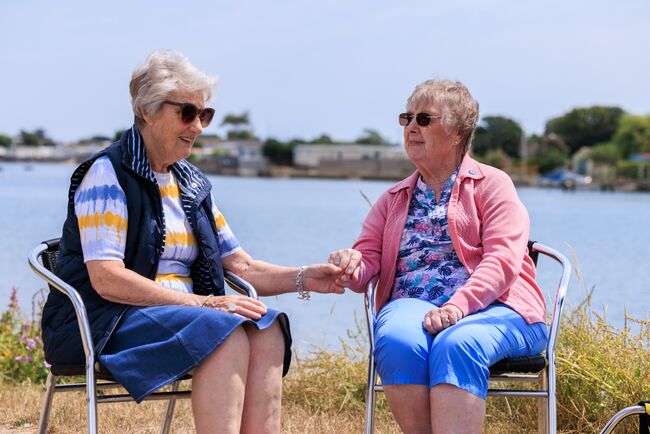 Maureen and Brenda holding hands sitting down in front of lake