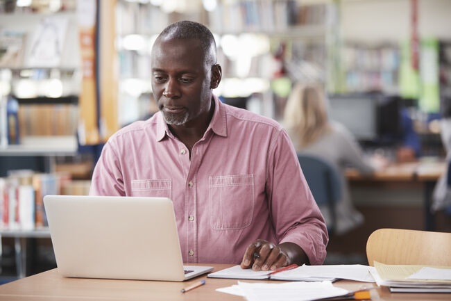 A stock image of a man sat in a library at a table, using a laptop with pens and papers on the table.