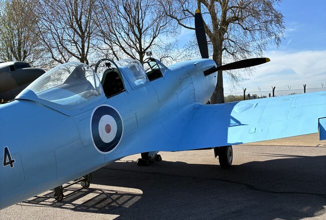 A blue Spitfire on the ground, from behind looking at the two, open cockpits